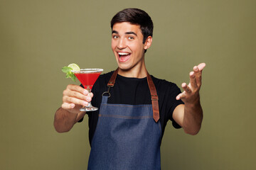 Young man barista bartender barman employee wear jeans apron black t-shirt work in coffee shop give glass of alcohol martini cocktail isolated on plain green background Small business startup concept