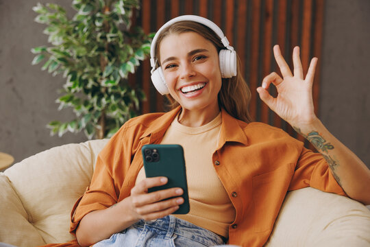 Young woman wear casual clothes listen music in headphones use mobile cell phone show ok sits in chair stay at home hotel flat rest relax spend free time in living room indoor. People lounge concept.