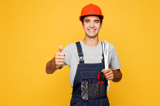 Fototapeta Young laborer handyman electrician man wear helmet overall carrying cables on shoulder show thumb up isolated on plain yellow background Instruments for renovation apartment room. Repair home concept