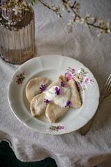 Dumplings with blueberry filling with addition of cream on a white plate, top view. A white cherry branches in blossom period with white flowers and fresh green leaves in glass vase on a white table