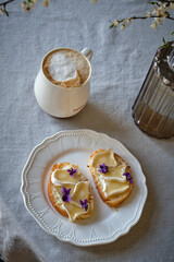 Warm Toast with cheese, camembert, wild violet and cappuccino on a white plate. A white cherry branches in blossom period with white flowers and fresh green leaves in glass vase on a white table