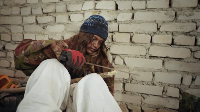 Unkept teen wearing dirty gloves and winter hat sharpens long piece of wood using cutlass while seated against brick wall in cold abandoned building, showing survival effort