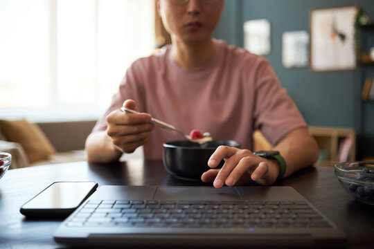 Asian young adult man sitting at table eating breakfast from bowl while using laptop and smartphone, focusing on multitasking with digital devices in modern home environment - Powered by Adobe