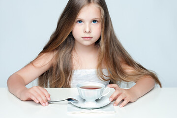 A serious young girl with long hair and blue eyes sits at a table with a cup of tea. Studio...