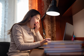A female student yawning from fatigue during her homework. A young girl sits at a desk with a laptop and yawns, showing her exhaustion from studying