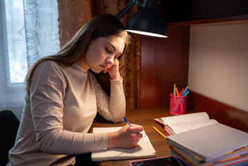 A tired female student preparing for exams at home. Young girl, tired and upset, sitting at the table and doing homework, homeschooling