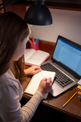 A female student is focused on writing in her notebook near a laptop. A young girl sits at a desk and carefully writes in a notebook, using her laptop for homeschooling