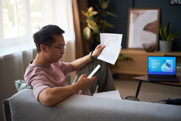 Asian young adult man sitting on sofa holding smartphone and examining financial documents, laptop with charts on table in background, working remotely in modern living room