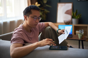 Asian young adult man sitting on sofa calculating finances using calculator and holding document, focused on managing personal budget with digital tablet and coffee cup on table nearby