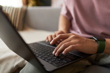 Young adult Asian man typing on laptop computer while sitting indoors, hands and partial torso...