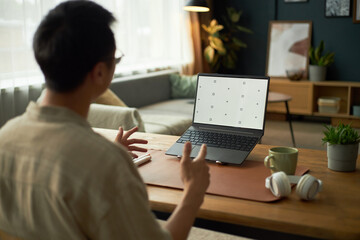 Asian young adult man gesturing while sitting at desk in home office, interacting with laptop...