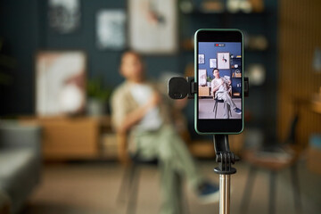 Young adult Caucasian man sitting on chair recording video with smartphone on tripod, holding cup and gesturing while looking toward device in modern indoor setting