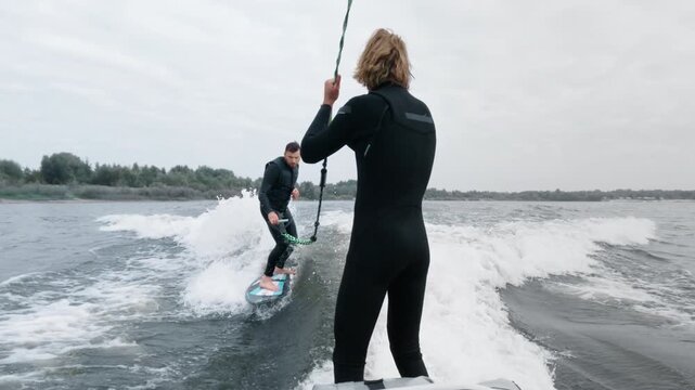 Two surfers engage in dynamic wakeboarding action on a calm water surface with cloudy skies