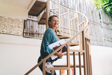 Fototapeta premium Caucasian woman walking upstairs with notes and tech gear reflects independence and control in flexible freelance life with mobile-first productivity mindset.