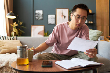 Asian young adult man sitting on sofa reviewing documents while holding paper in one hand and writing in notebook with pencil, glass pitcher and smartphone on table in living room