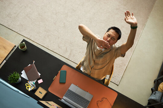 Asian middle aged man sitting at desk stretching arms and yawning while working with laptop and documents, smartphone and office supplies scattered on tabletop, top view