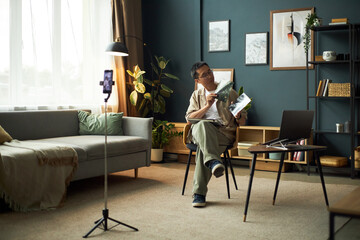 Fototapeta premium Asian middle aged man sitting on chair holding potted plant and gesturing, while recording video on smartphone tripod in modern living room with laptop on table