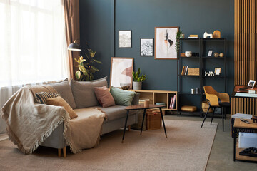 Modern living room featuring contemporary furniture, decorative plants, framed artwork on wall, open shelving with books and accessories, cozy sofa with cushions, coffee table, workspace setup