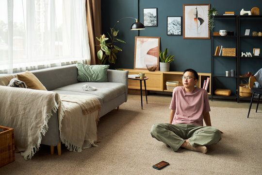Asian young adult man sitting cross legged on carpeted floor in modern living room looking upward with thoughtful expression, digital tablet lying nearby on floor - Powered by Adobe