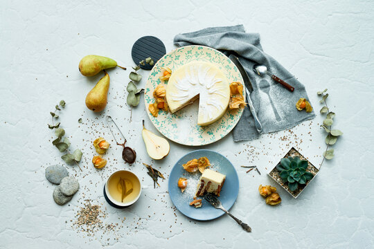 Overhead view showing sliced cheesecake on patterned plate surrounded by pears, cup of tea, physalis fruit, succulent plant, cutlery, linen napkin, and scattered seeds on surface
