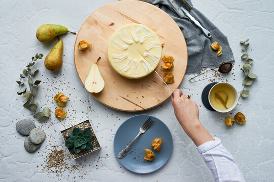 Caucasian young adult woman cutting cake with knife on wooden board, surrounded by pears, tea, succulents, and decorative elements, only hand and arm visible in frame
