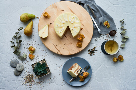Top view of cheesecake with decorative swirl pattern on wooden board surrounded by pears, physalis, cup of tea, succulent plant, seeds, and dried leaves on textured surface
