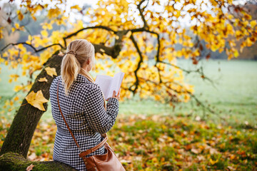 Woman is reading book in autumn park. Relaxation and mental wellbeing outdoors
