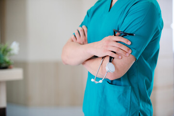 Close-up of unrecognizable doctor holding stethoscope while standing with arms crossed at medical clinic.