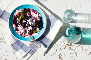 Bowl with yogurt topped with blueberries and mint leaves standing on striped cloth napkin, metal spoon resting inside bowl, glass and bottle of water placed nearby on textured surface
