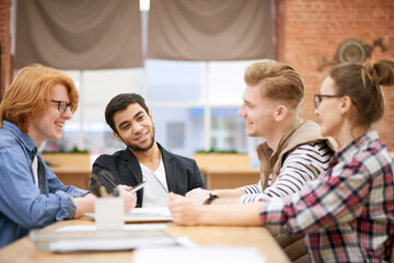 Meeting of creative team. Young enthusiastic male and female managers sitting at office desk, discussing last company achievements and smiling happily