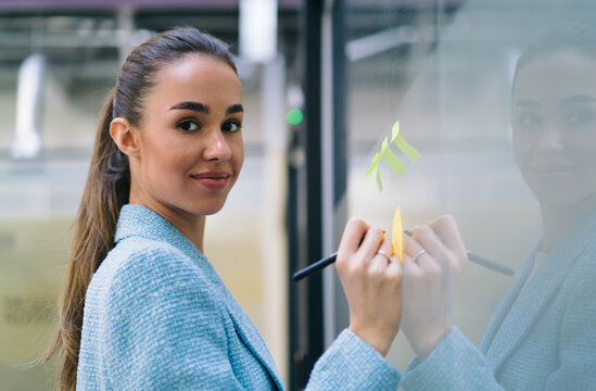 Smiling businesswoman looks at camera while placing notes on glass wall, expressing positive mindset, teamwork spirit and creative contribution in collaborative workspace. - Powered by Adobe