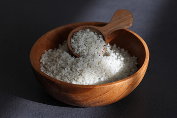 Dried coarse salt in wooden bowl with spoon in sun light on black table.