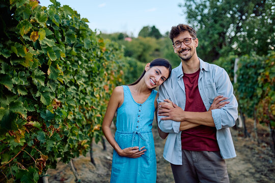 Expecting parents standing together in a vineyard looking at the camera. Woman holding her pregnant belly