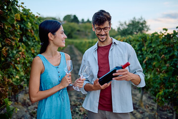 Happy couple holding wine bottle and glasses, standing among grapevines, enjoying a relaxing wine tasting experience
