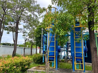 Industrial water treatment facility with large blue tanks and a network of pipes, surrounded by lush green trees and foliage in a natural outdoor setting.