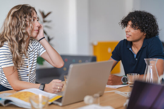 Two young people, a woman and a man, are having a discussion at a table. They are focused on their conversation, with a laptop and books nearby. This suggests a collaborative work or study session.