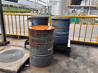 Three weathered industrial drums, two blue and one rusty, are placed in a service yard. The scene highlights themes of heavy industry, waste management, and manufacturing, with a factory building 