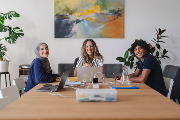 Three diverse young adults, two women and one man, sit at a table with laptops and notebooks, smiling at the camera. They are engaged in a collaborative work or study session.