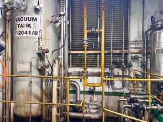 A wide-angle interior view of a complex industrial facility, featuring a large vacuum tank, intricate yellow handrails, and a dense network of pipes, valves, and gauges, conveying a sense of organized
