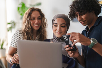 Three diverse young adults collaborate. A woman in a hijab and a man look at a smartphone, while another woman smiles at a laptop. They are sharing digital content.