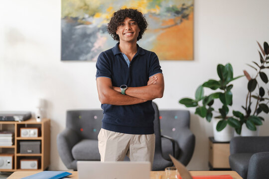 A smiling young man with curly hair stands confidently with crossed arms in a modern office. He wears a polo shirt and khaki pants, looking at the camera.