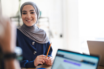 A smiling young woman in a hijab sits at a desk, holding a pencil. She is attentively participating in a discussion or meeting, with laptops nearby. Her expression is positive and engaged.