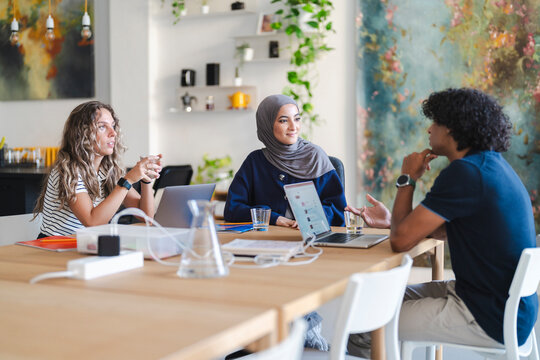 Three diverse young adults are collaborating at a modern office table. Two women and one man are engaged in a discussion, with laptops and drinks present.