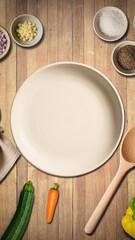 Vertical top-down view of a wooden kitchen table arranged with an empty plate, napkin, wooden spoon and fresh vegetables, capturing a warm and inviting home cooking atmosphere.