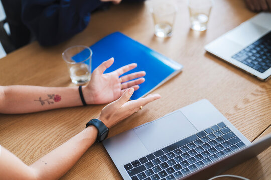 A person gestures with their hands during a business meeting or collaborative discussion. Laptops, a blue folder, and water glasses are on the wooden table. This shows teamwork and communication.