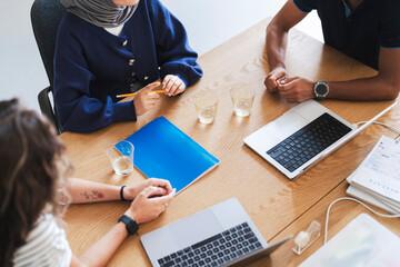 Three diverse individuals are collaborating at a wooden table. They are engaged in a meeting or study session, with laptops, a folder, and glasses present.
