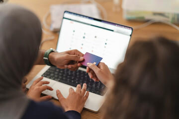 Two individuals exchange a credit card over a laptop. One person, wearing a hijab, hands the card to another. They are conducting an online financial transaction.