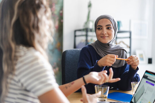 Two diverse women are engaged in a business meeting. The woman in a hijab listens intently while holding a pencil. They are collaborating in a modern office setting.
