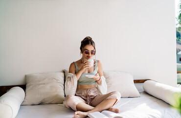 Obraz premium Stylish young woman drinking coffee on sofa with open book beside her. Blend of modern femininity, mindful habits and visual calm in natural tropical atmosphere.