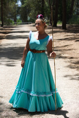 Fototapeta premium A beautiful young Spanish woman dressed in a traditional green flamenco dress and with a flower in her hair walks through the pine forests of Andalusia in the pilgrimage, leaning on a stick.